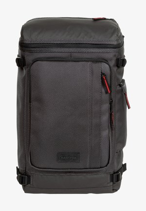 Mochila gris con un tejido texturizado, que cuenta con un gran compartimento principal, múltiples cremalleras en negro y rojo, y un asa resistente en la parte superior.