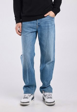 Person trägt hellblaue Jeans mit geradem Bein, weiße und graue Sneaker und ein schwarzes Langarmshirt, eine Hand in der Tasche.