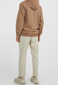Persona che indossa pantaloni beige, felpa color sabbia e sneakers beige, in piedi con le spalle rivolte verso uno sfondo chiaro e semplice.