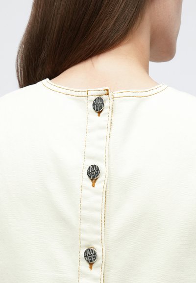 Vue de dos d'une femme portant une chemise crème avec trois boutons noirs centrés dans le dos, cousus avec un fil orange.