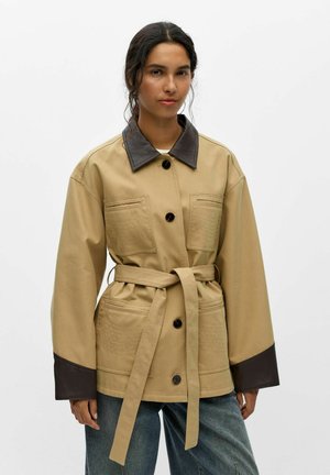 Donna che indossa una giacca beige con cintura, colletto e polsini neri, in piedi davanti a uno sfondo bianco uniforme.
