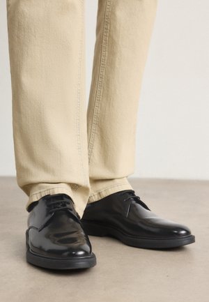 Chaussures habillées en cuir noir poli portées avec un pantalon beige à jambe droite, debout sur un sol lisse gris clair.