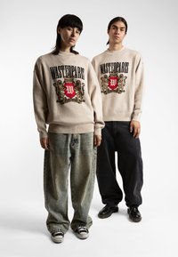Los suéteres beige presentan un emblema gráfico con un escudo rojo y leones. Se combinan con jeans holgados y descoloridos, y zapatos negros.