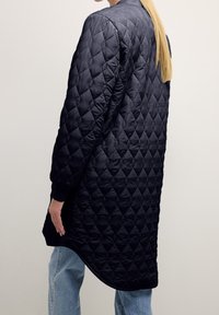 Giacca imbottita nera trapuntata con orlo lungo e arrotondato, polsini a costine e motivo a rombi. Tessuto morbido e materiale leggero.