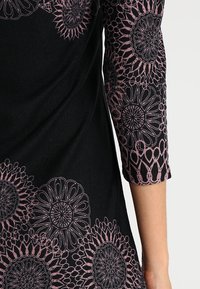 Svart tygplagg med trekvartslånga ärmar, prytt med rosa blommönster och intrikata design detaljer längs kanterna.