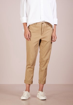 Personne portant un pantalon beige retroussé, des baskets blanches décontractées et une chemise blanche à manches longues avec les manches retroussées jusqu'aux coudes.
