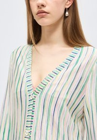 Γυναίκα που φοράει ζακέτα με V λαιμόκοψη με κάθετες παστέλ πράσινες, μπλε και μοβ ρίγες, κουμπωμένη με λευκά κουμπιά, και σκουλαρίκι σε σχήμα σταγόνας.