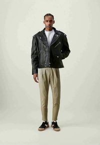 Jaqueta de cabedal preta de motard com detalhes em prata, bolsos com fecho e lapelas largas, combinada com calças beige claro e sapatilhas pretas.