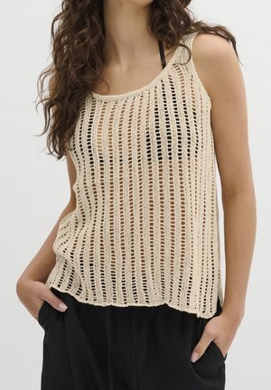 Femme portant un haut beige en crochet sans manches par-dessus un vêtement noir, mains dans les poches, sur un fond blanc uni.