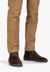 Hnědé semišové kotníkové boty s plochou gumovou podrážkou, vybavené šňůrkami a hladkým povrchem, nošeny s khaki kalhotami.