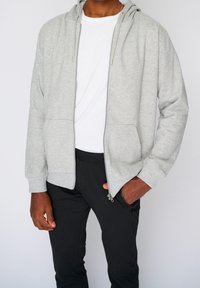 Felpa con zip grigia chiara, dotata di cappuccio con lacci, due tasche frontali e polsini a coste, indossata sopra una maglia bianca e abbinata a pantaloni neri.