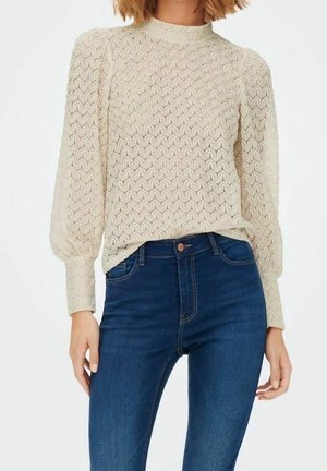Mujer que lleva un suéter de punto beige de manga larga con un patrón hexagonal y vaqueros azul oscuro de talle alto sobre un fondo liso.