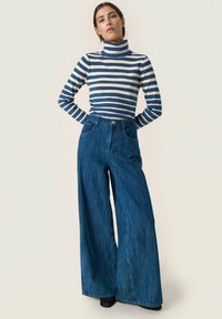 Prugasti turtleneck top u plavoj i krem boji, upareni s trapericama širokog kroja s visokom strukom u tamnom pranju. Oba artikla imaju minimalne detalje.