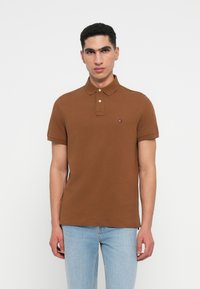 Καφέ πόλο μπλουζάκι από βαμβάκι, με γιακά, πλάκα με δύο κουμπιά και ένα μικρό λογότυπο στο στήθος. Κοντά μανίκια και ευθύ τελείωμα.