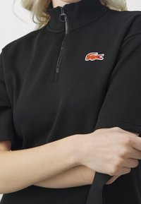 Czarny polo z zamkiem na ćwierć z miękką fakturą, z czerwonym i białym haftowanym logo krokodyla po lewej stronie klatki piersiowej. Krótkie rękawy.