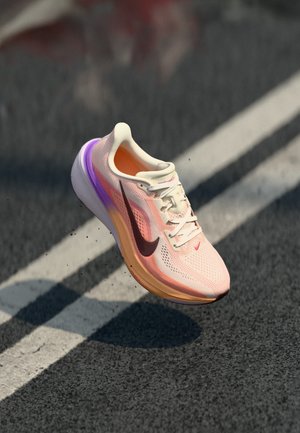 Lichtroze en oranje Nike hardloopschoen met paarse accenten zwevend boven asfalt en witte wegmarkeringen.