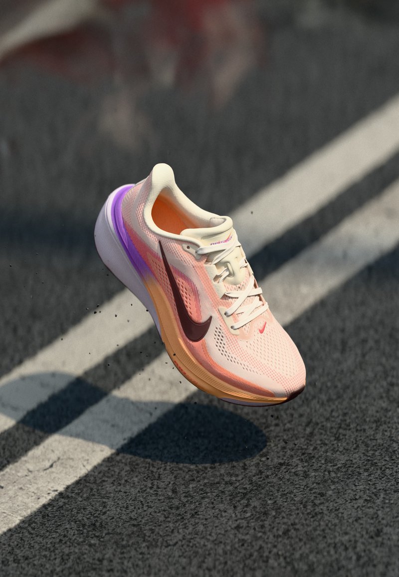 Chaussure de course Nike rose clair et orange avec des accents violets, flottant au-dessus de l'asphalte et des lignes blanches de la route.