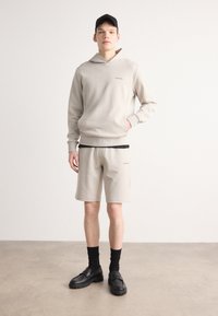 Sweat à capuche beige et shorts assortis, fabriqués en tissu doux. Détails noirs sur les shorts. Modèle portant des chaussures noires et des chaussettes.