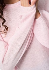 Close-up of person wearing soft pink knitted sweater, with hand gently touching the sleeve near the face, showing curly hair strand.