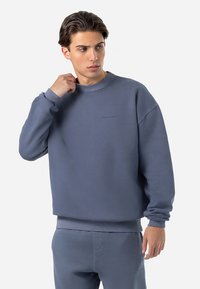 Blå sweatshirt med rund halsudskæring, blød tekstur og ribbede manchetter. Har et subtilt præget logo på brystet. Koordineret med matchende bukser.