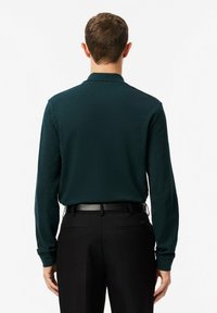 Dunkelgrünes Langarmpolohemd mit strukturiertem Finish und geradem Kragen, kombiniert mit schwarzen Hosen und einem Gürtel.