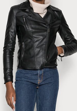 Giacca da motociclista nera in pelle con un design strutturato, tasche con zip e una texture liscia, abbinata a un dolcevita grigio chiaro e jeans.