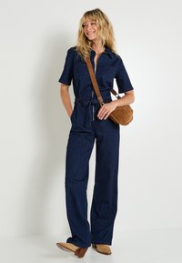 Mujer vestida con un mono de mezclilla azul oscuro de mangas cortas, botas de tacón de ante marrón y un bolso cruzado marrón, sonriendo de lado.