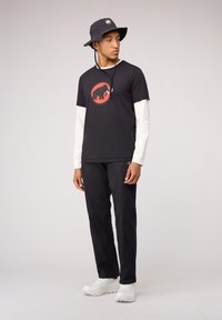 Zwarte katoenen t-shirt met een rode olifantgrafiek, gelaagd over een witte lange mouw. Gecombineerd met een zwarte broek en witte sneakers. Zwarte bucket hat.