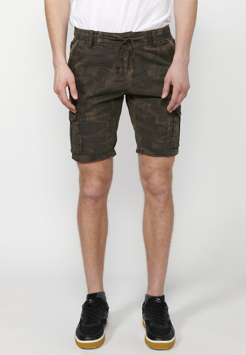 Tarnfarben Cargo-Shorts aus dunkelgrünem Stoff, mit elastischem Bund und Kordelzug, sichtbaren Nähten und seitlichen Taschen.