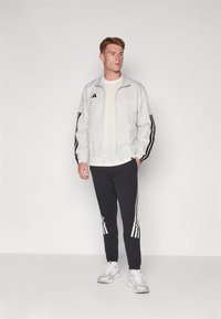 Giacca zip bianca con motivo e strisce nere ai lati, abbinata a pantaloni della tuta neri con strisce bianche sui lati e sneakers bianche.