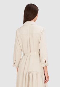 Beżowa sukienka koszulowa z paskiem, długimi rękawami, zaokrąglonym kołnierzykiem i marszczonym tylnym dołem. Materiał wydaje się lekki i ma gładką fakturę.