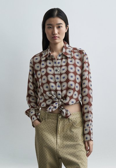 Femme aux cheveux longs et bruns porte un chemisier marron noué orné de marguerites blanches et un pantalon beige à petits motifs en points, debout devant un fond uni.