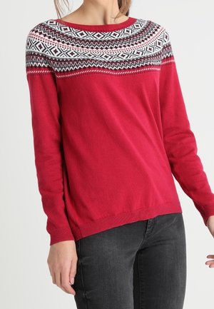 Czerwony sweter z długim rękawem z białym, czarnym i różowym wzorem Fair Isle na górnej części klatki piersiowej i ramionach, noszony z ciemnymi dżinsami.