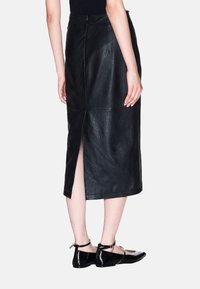 Gonna midi in pelle nera con spacco posteriore, finitura strutturata e dettagli con cuciture. Abbinata a ballerine nere lucide con cinturino alla caviglia.