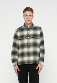Camisa de franela en tonos apagados de verde y crema, con un patrón a cuadros, cierre frontal de botones y dos bolsillos en el pecho.