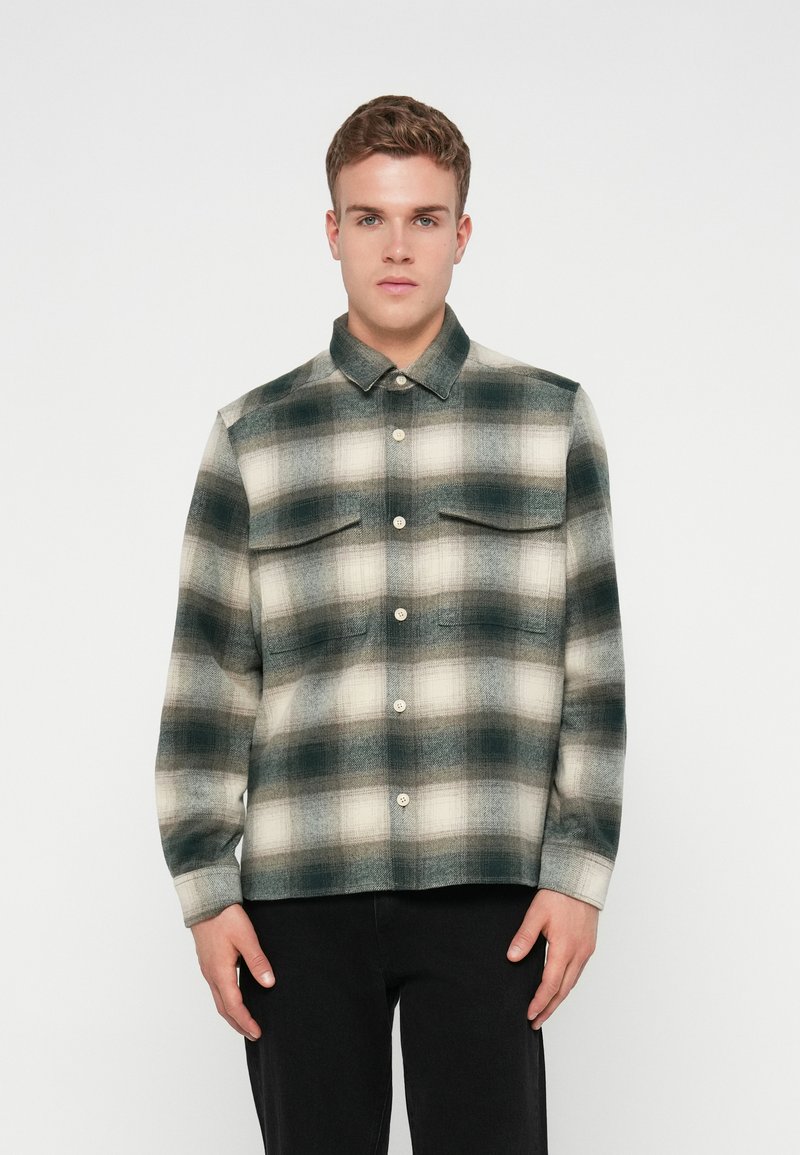 Camisa de franela en tonos apagados de verde y crema, con un patrón a cuadros, cierre frontal de botones y dos bolsillos en el pecho.