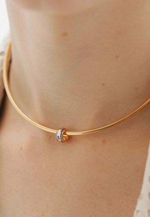 Collar de tono dorado con un diseño redondo y liso que presenta un anillo de acento azul y blanco grabado con la letra "B".