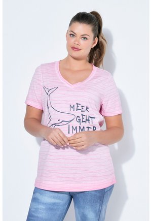 Kvinde med hestehale iført en lyserød, stribet V-hals T-shirt med en hval-grafik og tysk tekst, kombineret med blå jeans, mod en hvid baggrund.