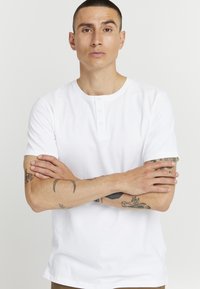 Balta īsām piedurknēm henley krekls, izgatavots no kokvilnas, ar apaļu kakla izgriezumu un trīs pogu aizdari. Gluda tekstūra, minimālistisks dizains.