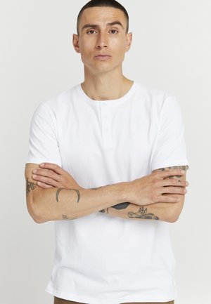 Camiseta henley blanca de manga corta hecha de algodón, con cuello redondo y tapeta de tres botones. Textura suave, diseño minimalista.