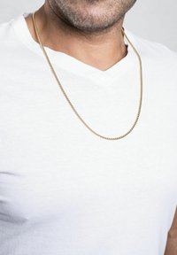 Gul kædedesign med en glat, rund form, hængende over en hvid V-hals t-shirt, der fremhæver dens metalliske glans og reflekterende overflade.