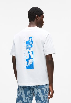 Férfi, fehér pólóban, hátul kék grafikával és "Karl Lagerfeld Jeans" felirattal, kék mintás farmer rövidnadrággal párosítva.