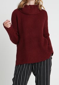 Bordowy sweter o prążkowanej teksturze, z oversize'owym kołnierzem i długimi rękawami. Połączony z czarno-białymi spodniami w paski.