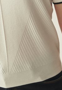 Haut en maille côtelée beige clair avec motifs verticaux texturés, ourlet en maille uni et accents noirs contrastants sur les manches.