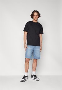 Sort bomulds t-shirt, blå denim shorts og sorte og hvide sneakers. Model står foran en ensfarvet hvid baggrund.