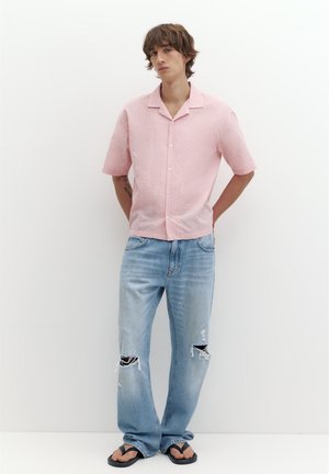 Giovane uomo con capelli castani che indossa una camicia rosa chiaro a maniche corte con bottoni, jeans azzurri strappati e infradito nere, in piedi contro un muro bianco.