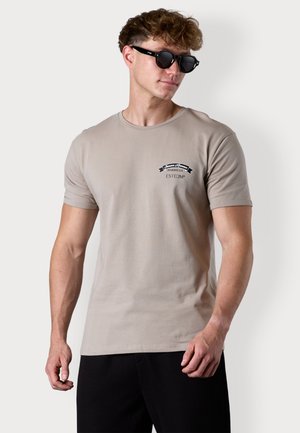 Hellbeige Baumwoll-T-Shirt mit kurzen Ärmeln, mit einem schwarzen Grafik- und Textdruck auf der Brust. Modell trägt eine dunkle Sonnenbrille.