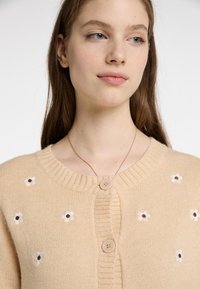 Beige gestrickte Strickjacke mit einem gerippten Ausschnitt, verziert mit gestickten weißen Blumen mit braunen Zentren und zwei vorderen Knöpfen.