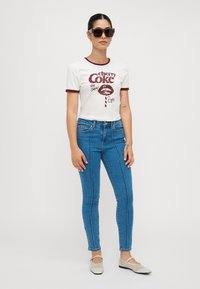 Bela skrajšana majica s temno rdečimi akcenti, ki prikazuje grafični dizajn Cherry Coke. V kombinaciji z modrimi skinny džinsom in svetlimi balerinkami.