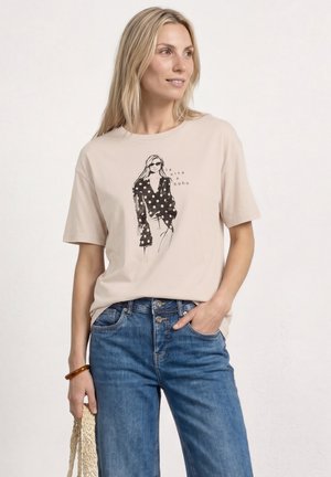 Mujer con camiseta beige con ilustración de moda de lunares negros y texto "la vita è boho", combinada con jeans azules, sosteniendo una bolsa tejida.